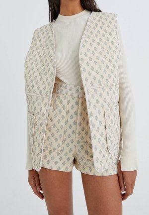 Femme portant un haut à manches longues en maille côtelée crème, un gilet matelassé assorti et un short à petit imprimé floral avec de grandes poches avant.