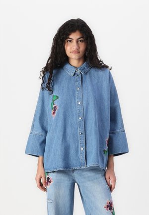 Lichtblauwe spijkerblouse met kraag, voorzien van bloemenborduurwerk in roze en groen aan de voorkant en losvallende, klokvormige mouwen.