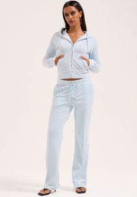 Helles blaues Trainingsanzug aus weichem Stoff. Verfügt über eine Kapuzenjacke mit Reißverschluss und Taschen sowie eine Hose mit Kordelzug und Seitentaschen. Glatte Textur.