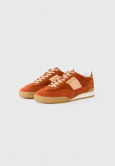 Un paio di sneakers basse in arancione ruggine con lacci beige, pannelli in tessuto testurizzato e suole in gomma su uno sfondo neutro.