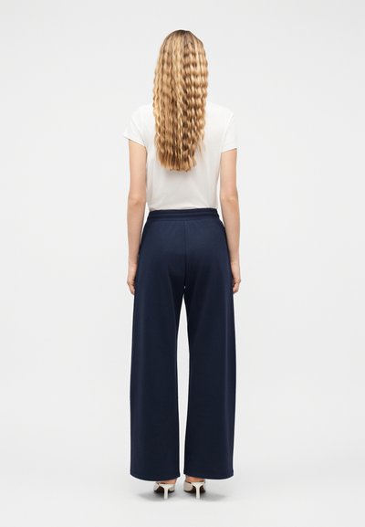 Γυναίκα με μακριά κυματιστά ξανθά μαλλιά στέκεται με την πλάτη, φορώντας λευκό κοντομάνικο τοπ, ναυτικό παντελόνι με φαρδιά πόδια και λευκά παπούτσια με τακούνι.