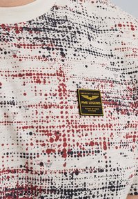 Soveblå t-shirt i bomuld med en cremefarvet baggrund, prydet med et prikket mønster i rød og marineblå. Indeholder et sort og guld mærke på brystet.