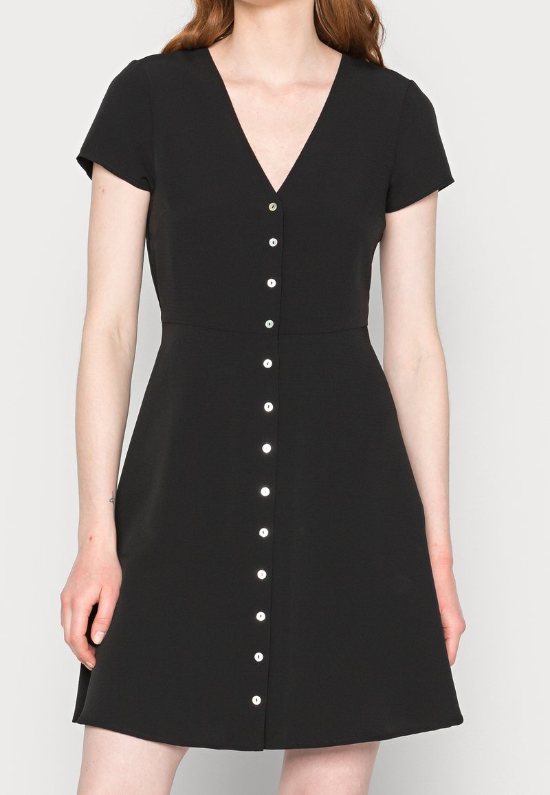 Femme portant une robe noire à manches courtes avec un col en V et une rangée de boutons blancs au centre devant.
