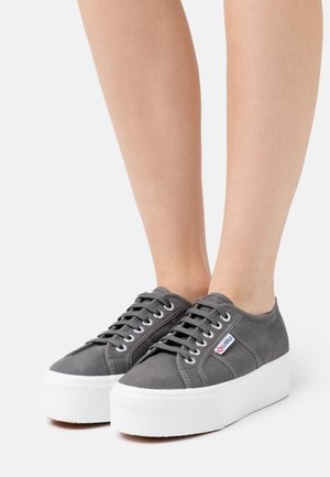 Personne portant des baskets compensées en toile grise avec des semelles épaisses blanches et des lacets noirs, debout devant un fond clair uni.