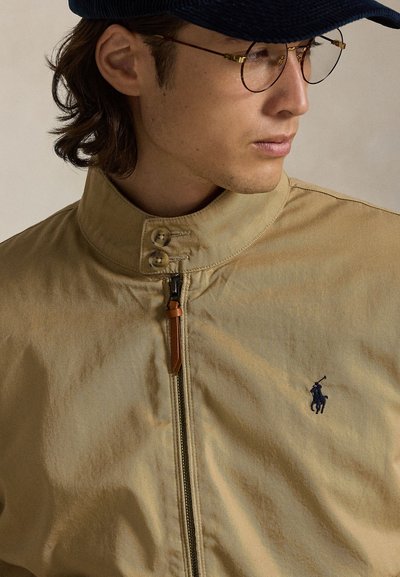 Noor mees, kes kannab beežika luku ja kahe nööbiga kraega jopet, väikest tikitud polo mängija logo, mereväe sinist nokamütsi ja ümarate kuldsete äärega prille.