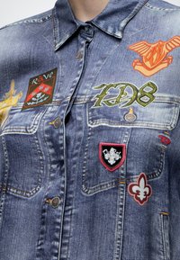 Veste en jean bleu avec plusieurs écussons, dont un aigle orange, des symboles "7D8," fleur-de-lis verts et un dessin de bouclier rouge et noir.