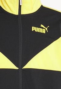 Veste zippée noire et jaune avec logo Puma jaune sur le côté droit de la poitrine, avec un design en blocs de couleur et un col montant.