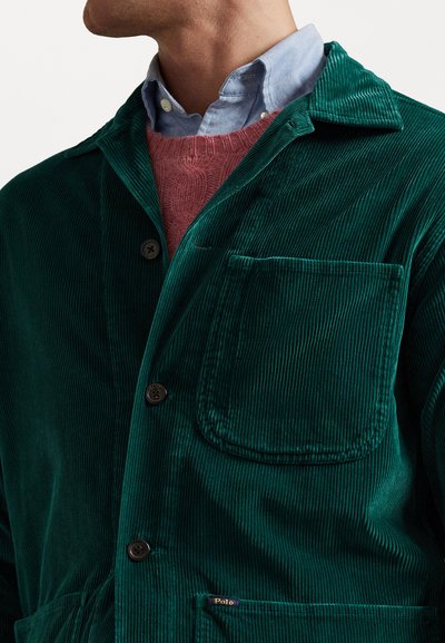 Green corduroy jacket with a notched collar, front pocket, and black buttons. Worn over a pink sweater and a light blue button-up shirt.
