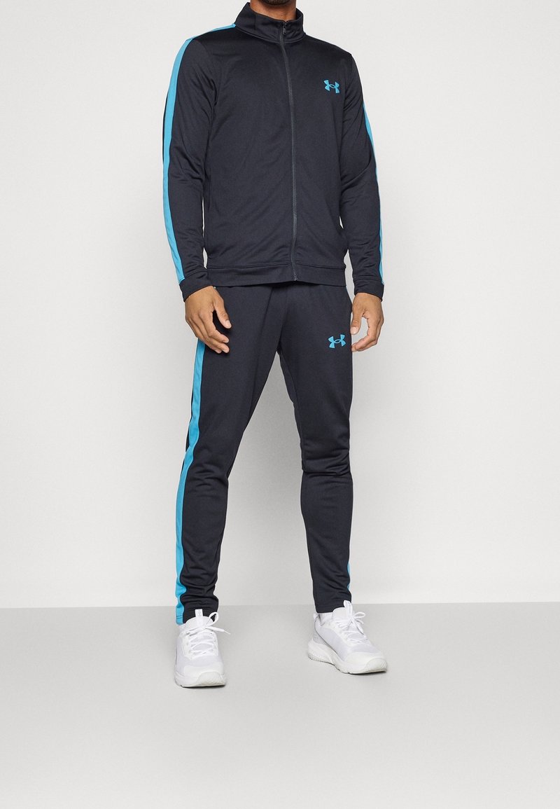 Homme portant un survêtement noir avec des bandes bleu clair aux manches et aux jambes, et des chaussures de sport blanches, debout devant un fond uni.