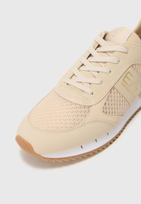 Beige sneakers med nettpaneler, rund tå og snøresystem. Har gummisåle og subtil merkevare på siden.