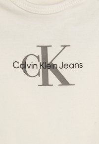 Tricou din bumbac crem, cu un logo gri "CK" și textul "Calvin Klein Jeans" dedesubt. Design clasic cu guler rotund și textură netedă.