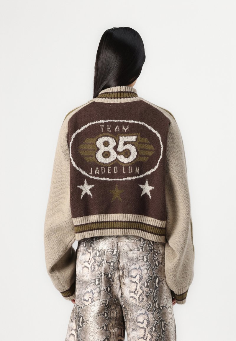 Knit jacket featuring a brown upper with "TEAM 85" in white, beige sleeves, ribbed hem, and star details. Worn with patterned metallic pants.