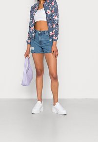 Bomber jacket floreale su sfondo blu, top corto bianco, pantaloni shorts in denim strappati e borsa viola. Il modello indossa sneakers bianche con plateau.