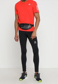 Červené sportovní tričko se třemi bílými pruhy, černé legíny a černá ledvinka s oranžovými akcenty. Černé a neonově zelen�é boty.