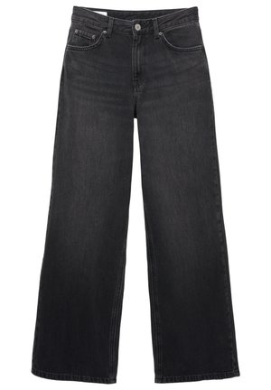 Ung kvinde står barfodet iført en hvid tanktop og højtaljede, vidde sorte jeans foran en ensartet hvid baggrund.