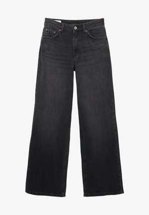 Ung kvinde står barfodet iført en hvid tanktop og højtaljede, vidde sorte jeans foran en ensartet hvid baggrund.