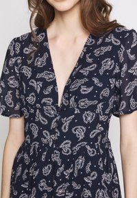 Laivansinis mekko valkoisella paisley-kuviolla, syvä V-aukko, lyhyet hihat ja napitus edessä. Hame on flare-mallinen ja kerätty vyötäröltä.