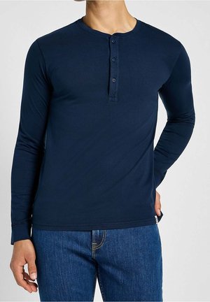 Mann trägt ein marineblaues Henley-Oberteil mit langen Ärmeln und drei Knöpfen, kombiniert mit blauen Jeans, Hand auf der Hüfte ruhend.