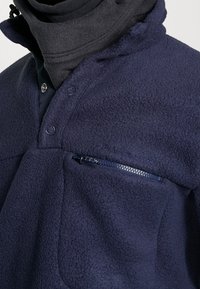 Sudadera polar azul marino con cierre de medio botón, bolsillo en el pecho con cremallera y tejido texturizado. Presenta costuras con la marca y diseño de cuello estratificado.