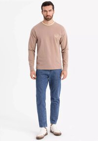 Pitkähihaisessa beige T-paidassa on pyöreä kaula-aukko, jossa on pieni logo-yksityiskohta. Yhdistettyinä sinisiin farkkuihin ja valko-ruusukkeisiin lenkkareihin.