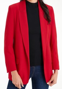 Rød blazer med afslappet pasform, hakket revers og forlommer, kombineret med en sort ribbet rullekrave og mørke jeans.