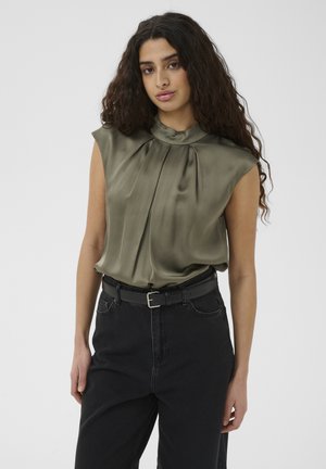 Jeune femme aux longs cheveux bouclés portant un chemisier sans manches en satin vert olive, rentré dans un jean noir taille haute avec une ceinture noire.