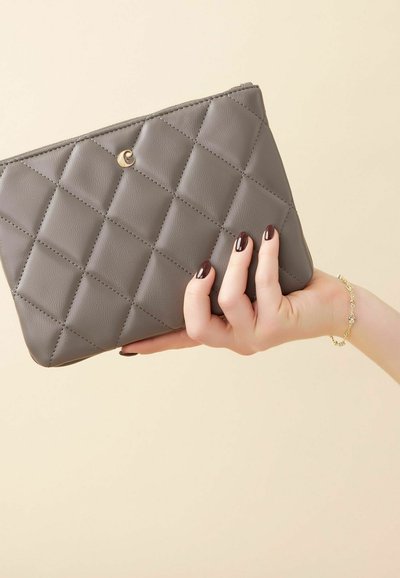Grijze gewatteerde synthetische leren pouch met een ritssluiting, met een ronde gouden accent en vastgehouden door een hand met donkere nagellak.