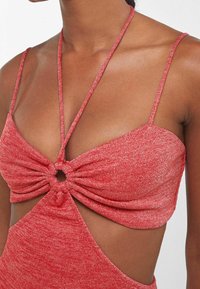 Punainen neulepaita, jossa on ohuet hihnat, kierretty etuosa ja pyöreä aukko, sekä sileä, kiiltävä pinta.