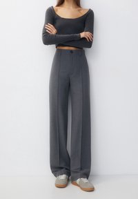 Vrouw staat met gekruiste armen, gekleed in een donkergrijze lange mouw crop top, hooggetailleerde grijze broek met wijde pijpen en beige sneakers.