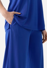 Modrý top a odpovídající kalhoty vyrobené z hladkého materiálu. Top má boční kapsu; kalhoty jsou volné a mají rovný střih.