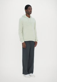 Hoodie verde chiaro con logo "EA7" in rilievo, tasca a marsupio e cappuccio con cordino, abbinato a pantaloni grigi scuri a vestibilità morbida e sneakers bianche.