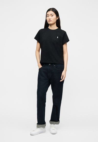 Νεαρό άτομο στέκεται με τα χέρια στις τσέπες φορώντας μαύρο κοντομάνικο μπλουζάκι, σκούρο τζιν με γυρισμένα μπατζάκια και λευκά αθλητικά παπούτσια σε λευκό φόντο.