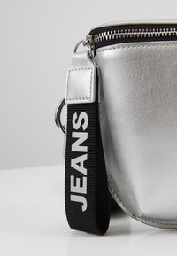 Borsa argentata con una finitura texturizzata, dotata di una tracolla nera con l'etichetta "JEANS" in bold lettere bianche. Chiusura con zip nella parte superiore.