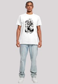 Hvit bomulls T-skjorte med svart grafisk design av en superhelt. Modellen har på seg lyseblå jeans og hvite sneakers.