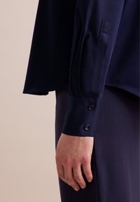 Granatowa bluzka o gładkiej teksturze, z mankietem wykończonym dwoma czarnymi guzikami oraz małym haftowanym kwiatowym detalem na mankiecie.