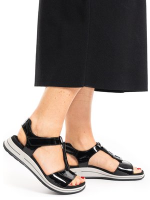 Füße in schwarzen, offen getragenen Sandalen aus Lackleder mit verstellbaren Riemen und weißen, gepolsterten Sohlen, kombiniert mit einem schwarzen Rock.