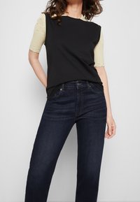 Černý top s krátkými, texturovanými žlutými rukávy; spárovaný s tmavě modrými džíny. Tkanina vypadá hladce a obepíná postavu, má jednoduchý design.