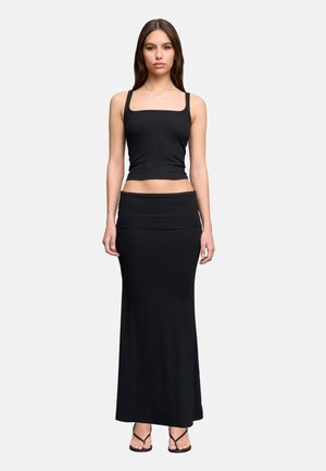 Junge Frau trägt ein schwarzes ärmelloses Crop-Top, langen schwarzen Rock und schwarze Sandalen, steht vor einem schlichten hellen Hintergrund.