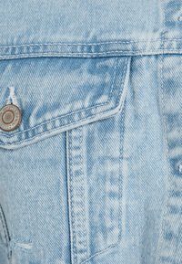 Ljusblå denimjacka med en silverknapp, synliga sömmar och en framficka med en distinkt spetsig klaff.