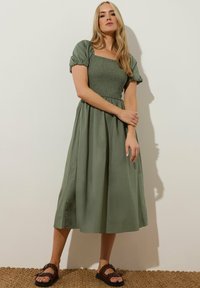 Olivgrünes Midikleid mit einem gerüschten Oberteil und Puffärmeln; leichtes Material, fließender Rock, getragen mit braunen, mit Nieten besetzten Riemchenschuhen.
