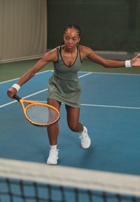 Vestido atlético sem mangas para mulheres em verde oliva, com um decote em V e silhueta ajustada. Jogadora balança uma raquete de tênis com um quadro laranja.