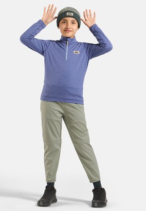 Child wearing blue long-sleeve top, olive pants, beanie, and black shoes, standing with hands raised beside head against white background.