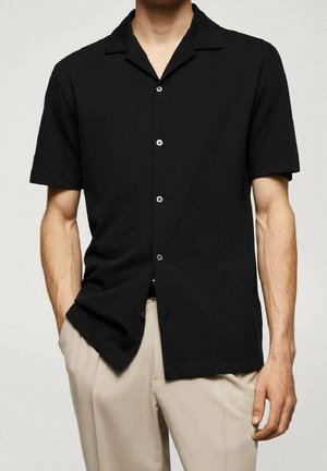 Homme portant une chemise noire à manches courtes avec col de camp, associée à un pantalon beige, les mains détendues le long du corps.