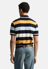 Mężczyzna w granatowej, żółtej, białej i jasnoniebieskiej w paski koszulce polo z krótkim rękawem, zestawionej z niebieskimi dżinsami i brązowym paskiem, widziany od tyłu.