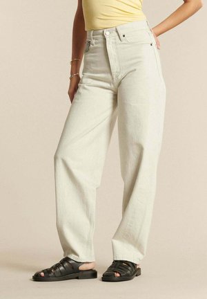 Osoba ubrana w kremowe, szerokie jeansy z wysokim stanem oraz czarne sandały z zakrytymi palcami, z jedną ręką na biodrze i widoczną żółtą bluzką.