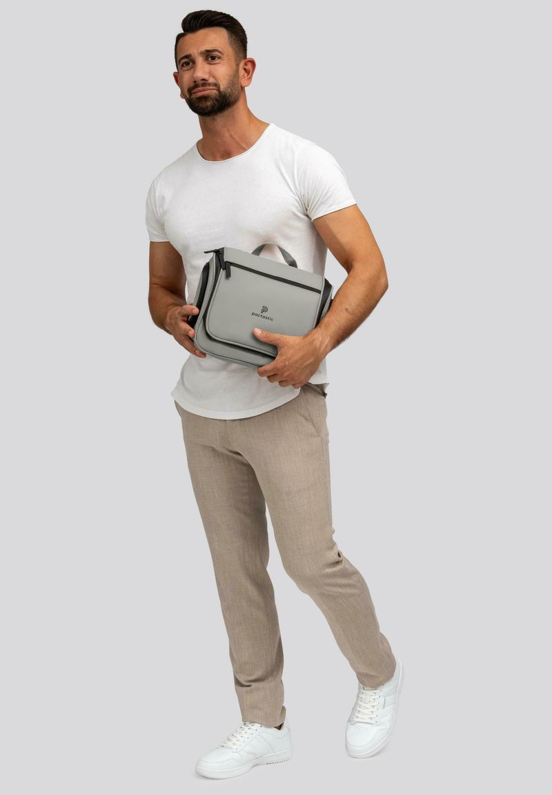 Borsa strutturata grigia con chiusura a zip e doppi manici. Materiale liscio, con un logo sottile. Il modello indossa una t-shirt bianca e pantaloni beige.
