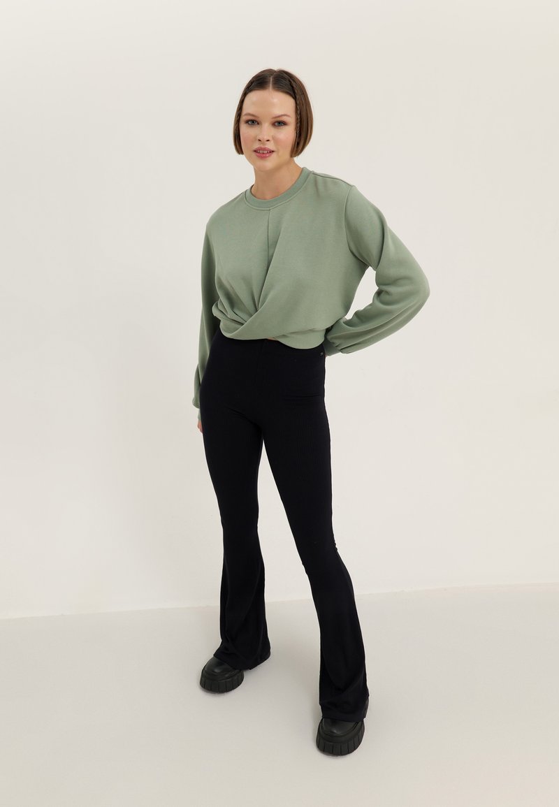 Top verde de manga larga con diseño de frente retorcido, combinado con pantalones acampanados negros. La modelo lleva zapatillas negras gruesas. Fondo neutral.