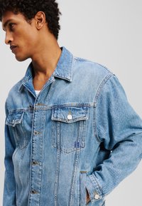 Ljusblå denimjacka med spetsig krage, två bröstfickor och silverknappar. Har synlig sömnad och en avslappnad passform.