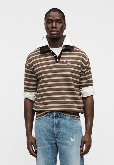 Καφέ και μπεζ ριγέ πόλο με λευκό γιακά πάνω από γκρι μπλούζα. Φοριέται με ανοιχτόχρωμα τζιν με κόκκινη κεντητική και σχέδιο πουλιού.