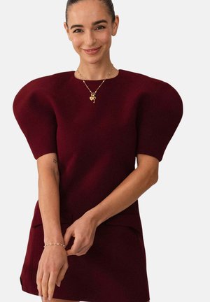 Frau lächelt, trägt ein burgunderrotes Kleid mit großen, aufgeblähten Ärmeln, eine goldene Halskette mit Schlüssel- und Herzanhängern sowie ein Perlenarmband.